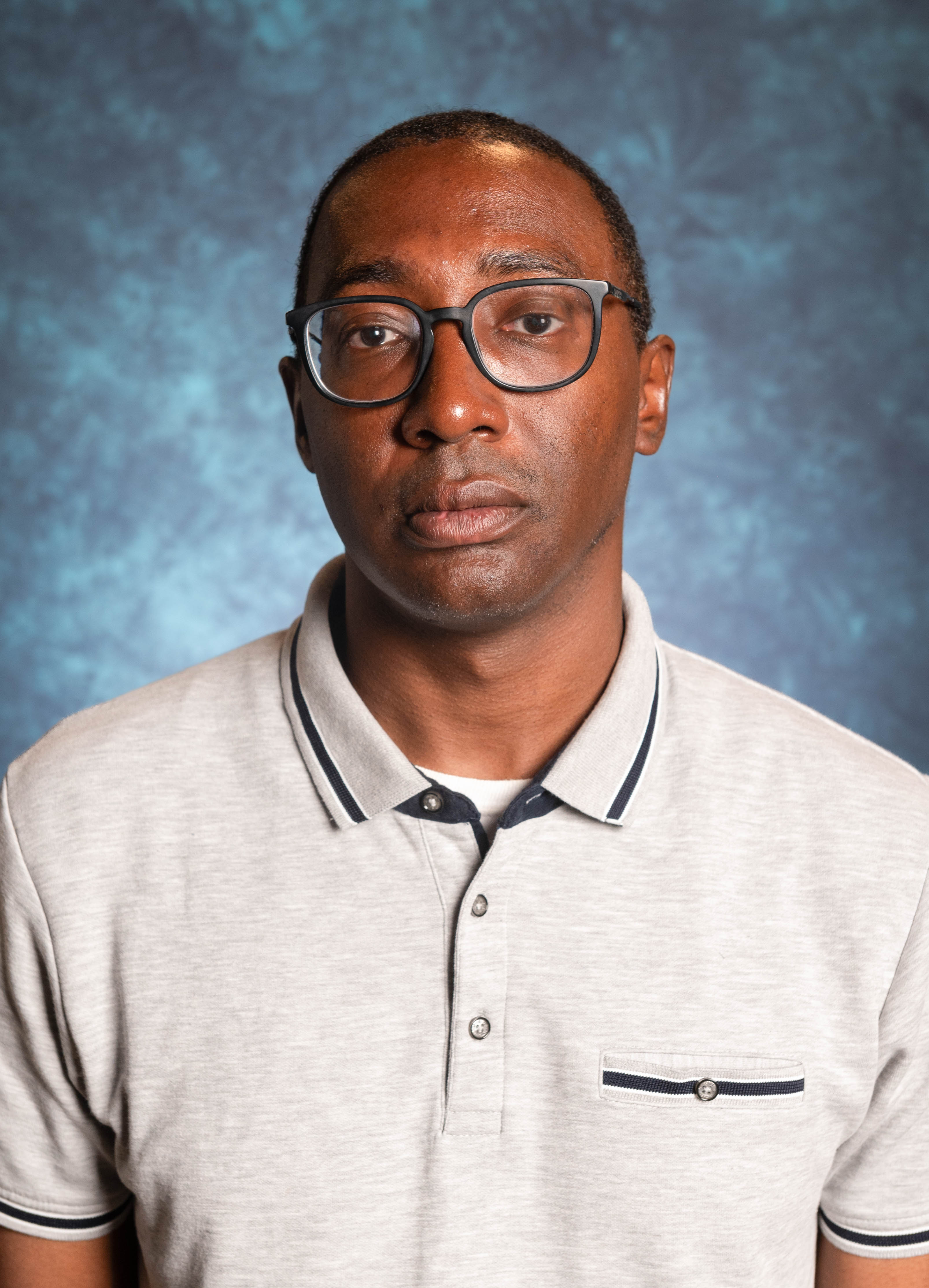 Picture of Cecil Rose, Director of Black Programs The image is a portrait of a person taken against a blue mottled background. He has a neutral expression and is wearing rectangular glasses with dark frames. His hair is short and he is dressed in a light gray polo shirt with a collar and short sleeves. The polo shirt features dark blue trim along the collar and sleeve edges, two buttons on the placket, and a small pocket on the left side of the chest with the same dark blue trim and a button. The background provides a soft contrast to the subject, allowing him to stand out clearly in the image.