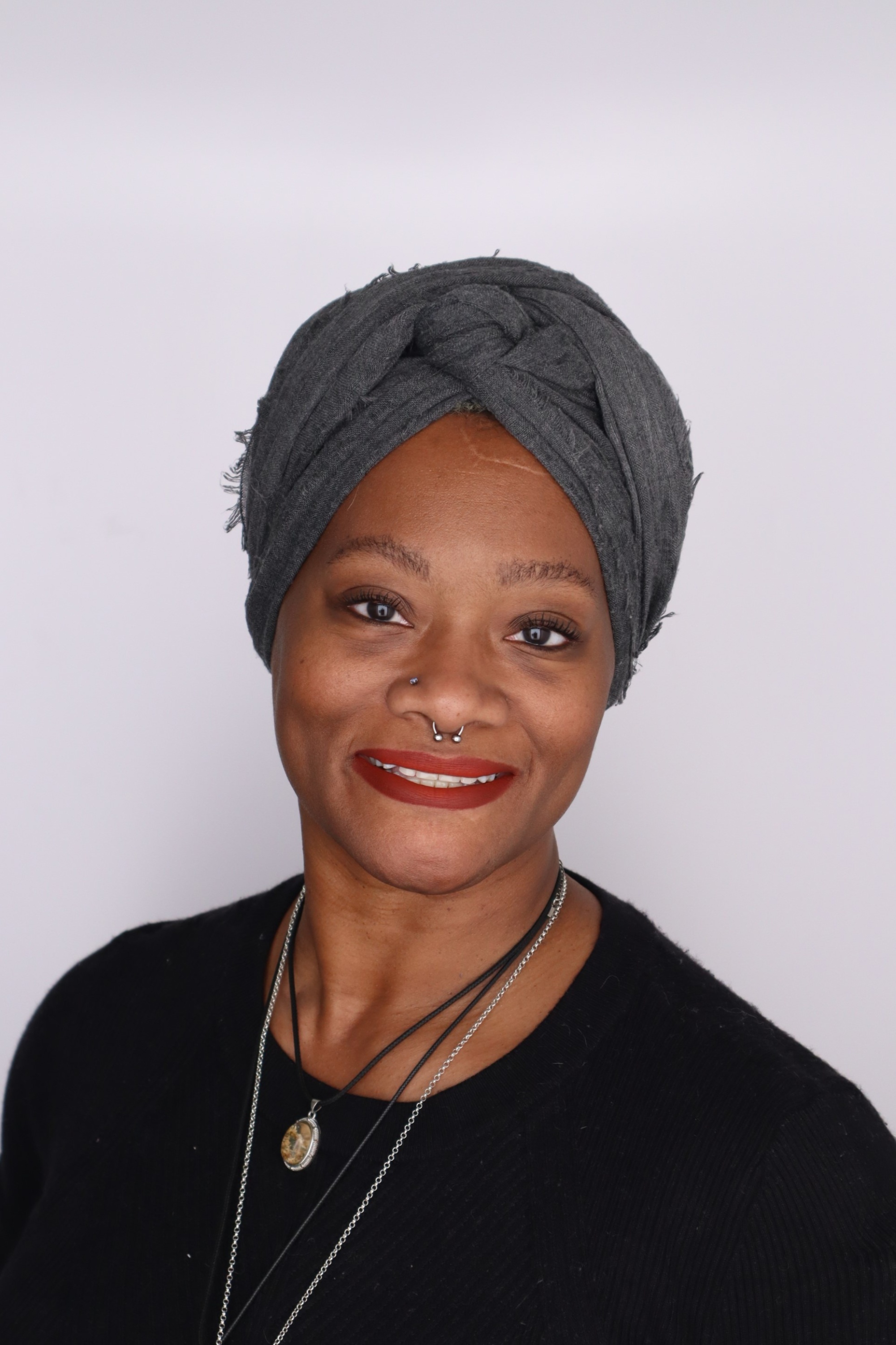 The image is a portrait of an individual against a plain, light-colored background. She is wearing a dark grey headwrap that covers her hair and is wrapped in a knot at the front. Natalie has light brown skin and is smiling. She is wearing a black top and several layered necklaces, one of which features a pendant with a circular design. She has a septum piercing and her makeup includes red lipstick and visible eyeliner. The overall lighting of the image is bright and even.