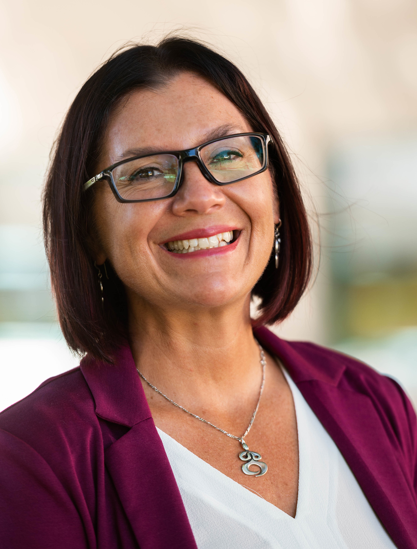 Picture of Teresa Maria Linda Scholz, Vice President, Equity, Inclusion, and Diversity  The image portrays a smiling person with shoulder-length dark hair, wearing rectangular glasses with thin, dark frames. She is dressed in a dark magenta blazer over a white top. Around her neck, she wears a silver necklace with a pendant that appears to be a stylized number eight or an infinity symbol. The background is softly blurred and indistinct, focusing attention on her. The lighting is bright and natural, providing a clear and well-lit image of her face and attire.