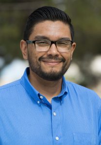 Picture of Joseph Duran, Program Coordinator of Chicano program. The image is a portrait of a man from the chest up. He is facing the camera and smiling slightly. The man has short, dark hair that's neatly combed back and sports a short beard and mustache. He is wearing dark-rimmed glasses and a bright blue button-up shirt with the top button undone. The background is blurred, indicating it might have been taken outdoors, with greenery and sky elements vaguely visible.
