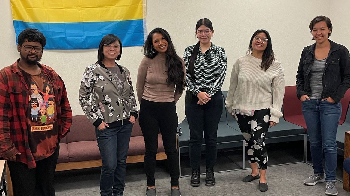 The image shows a group of six people standing in a row in a room. The background features a wall with a yellow and blue flag hanging on it. From left to right, the first person is wearing a red and black flannel shirt over a graphic T-shirt, the second person is wearing glasses and a floral jacket with jeans, the third person is wearing a beige long-sleeve top and black pants, the fourth person is wearing glasses and a black and white polka-dotted blouse with black pants, the fifth person is wearing glasses and a white sweater with floral pants, and the sixth person is wearing a black jacket over a grey shirt with jeans. There are maroon and white cushioned benches behind them.
